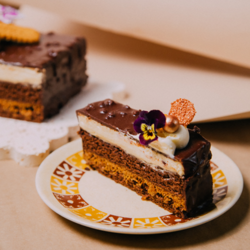 Slice of chocolate cake with cream and edible flower on a decorative plate.