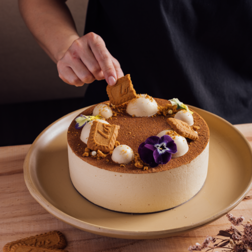 Person decorating cake with cookies and flowers on a wooden table.