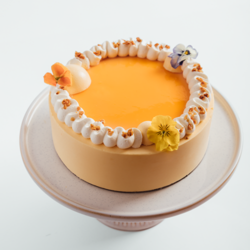 Elegant cake with orange glaze, cream, and flower decorations on a stand.