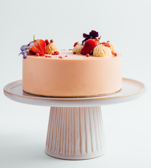 Peach-colored cake with strawberries and flowers on a white stand.