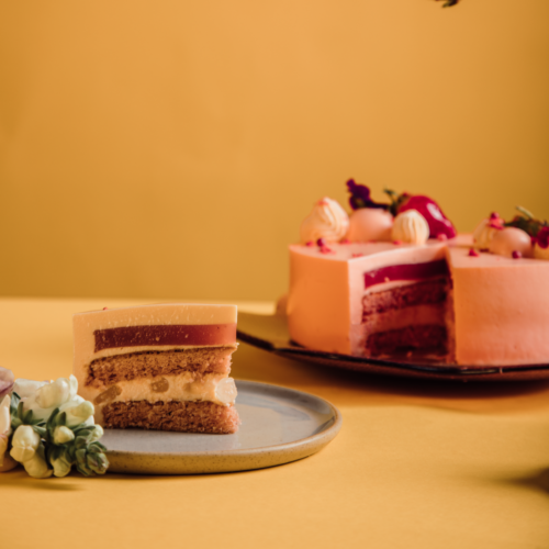 Slice of layered cake on plate, whole cake in background, yellow backdrop.