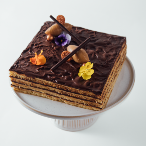 Layered chocolate cake with decorative flowers and chocolate sticks on a white cake stand.