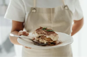 A Person in an apron holding a plate of homemade tiramisu