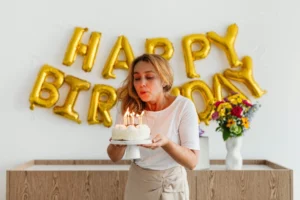 Woman blowing out candles on birthday cake, with "Happy Birthday" balloons behind.