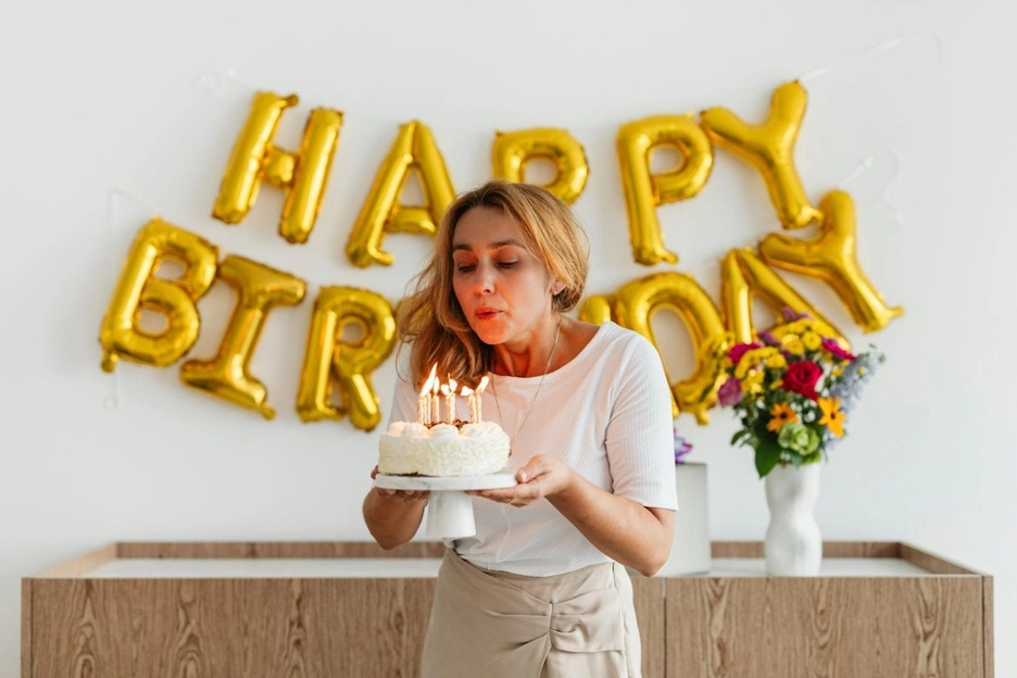 Woman blowing out candles on birthday cake, with "Happy Birthday" balloons behind.