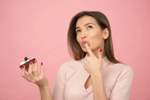 Woman pondering while holding a cupcake against a pink background.