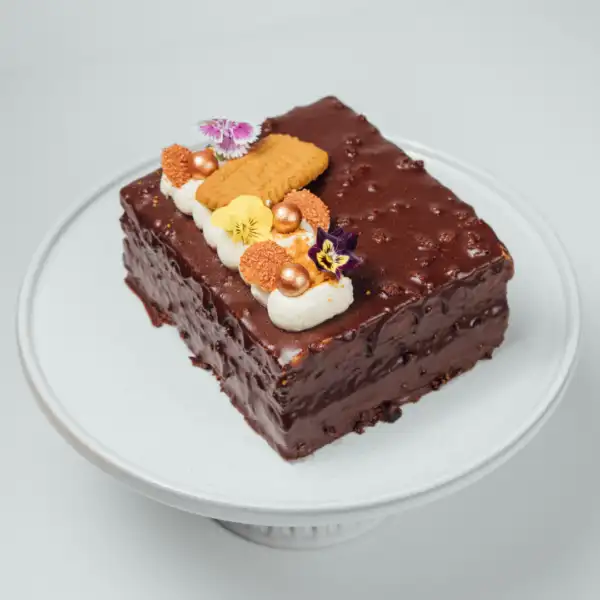 A chocolate cake with a glossy, textured chocolate coating, decorated with whipped cream, a biscuit, flowers, and gold-colored spheres