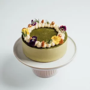 A green matcha cake decorated with cream, edible flowers, and gold pearls on a white cake stand.