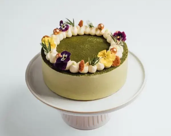 A green matcha cake decorated with cream, edible flowers, and gold pearls on a white cake stand.