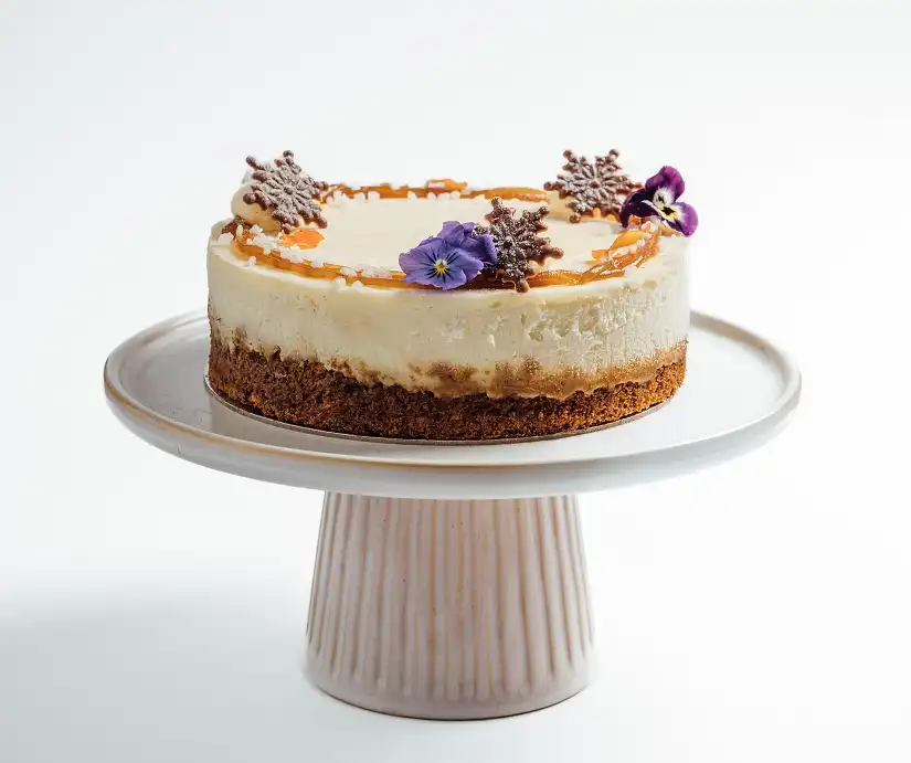  A round layered cake with cream topping, decorated with chocolate pieces and purple flowers, placed on a white cake stand.