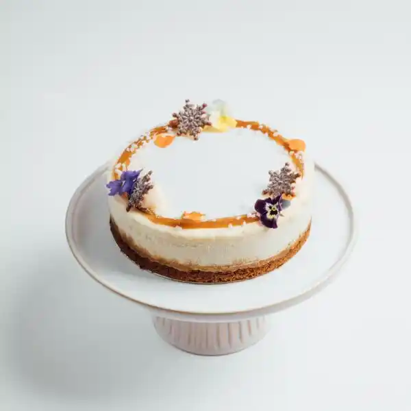 Decorative cheesecake topped with caramel drizzle, flowers, and chocolate accents, displayed on a white cake stand